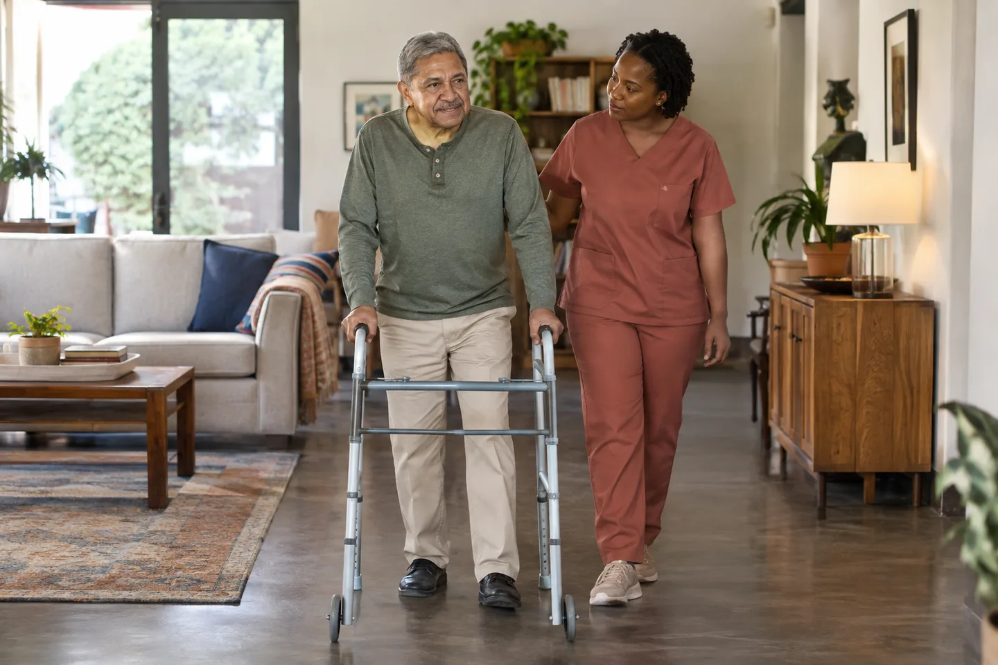 Illustrative scene: An older man stands with a walker while a female caregiver in terracotta scrubs stands attentively beside him